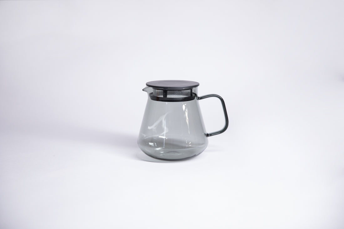 Transparent clear grey colored tulip shaped tea or coffee server with glass handle and fluted spout and black lid with a built in strainer. Set on white background