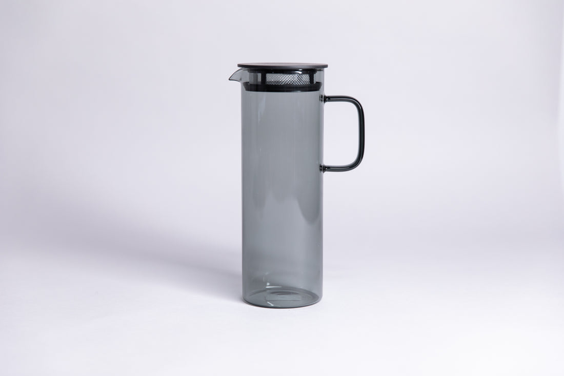 Cylindrical transparent gray colored glass pitcher with a fluted spout and rectangular handle with a black colored filtered lid. set on a a white background. 
