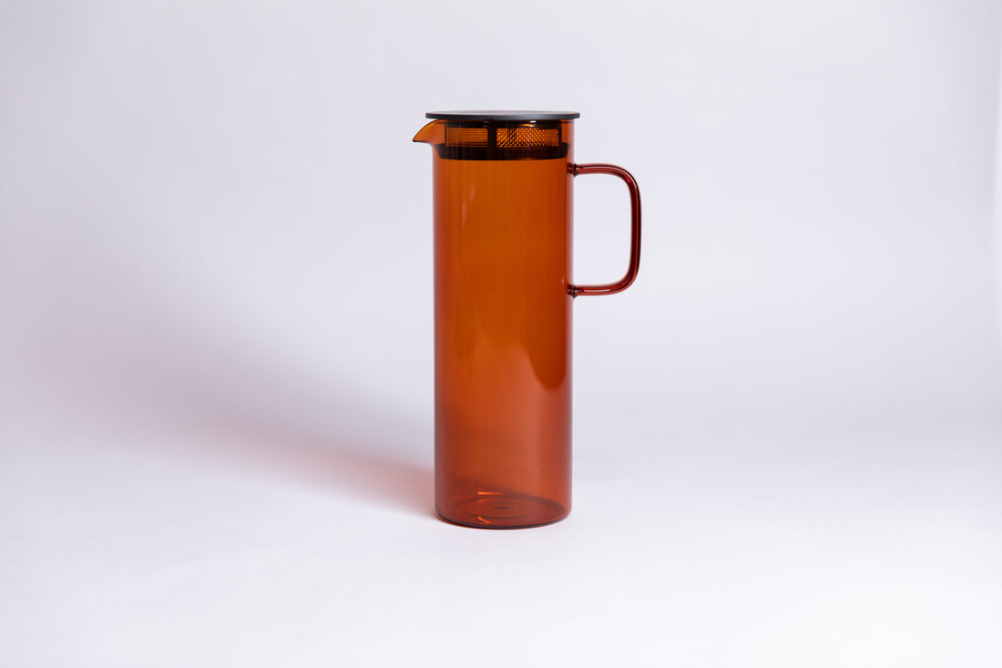 Cylindrical transparent amber colored glass pitcher with a fluted spout and rectangular handle with a black colored filtered lid. set on a a white background. 