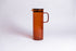 Cylindrical transparent amber colored glass pitcher with a fluted spout and rectangular handle with a black colored filtered lid. set on a a white background. 