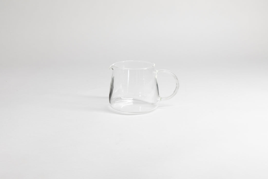 Round glass server with glass handle and fluted spout. On a light grey background.