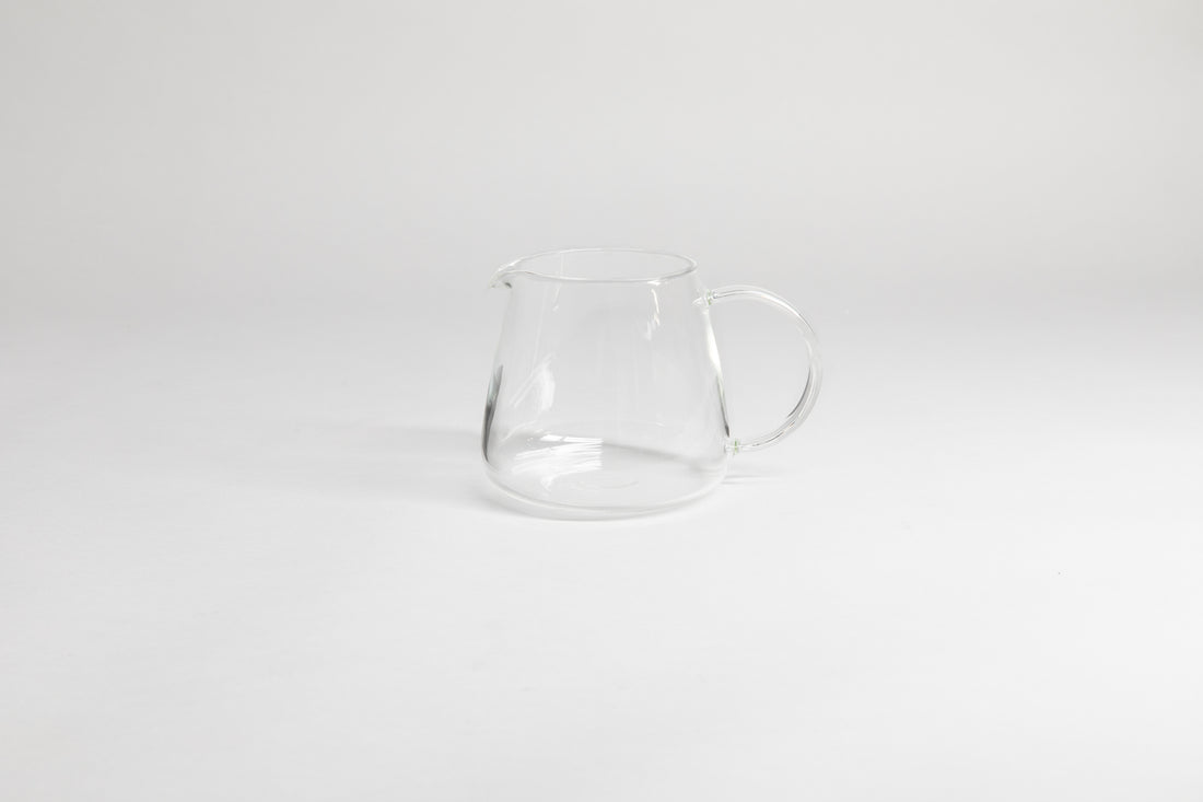 Round glass server with glass handle and fluted spout. On a light grey background.