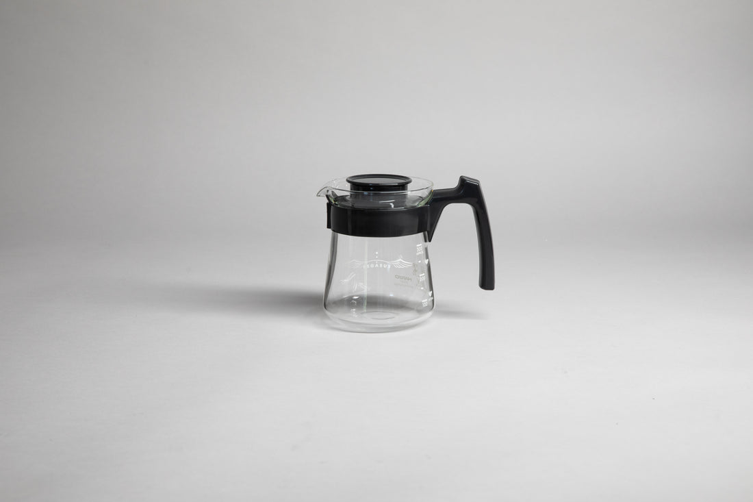 Glass coffee server with white text and level markings with black hard plastic handle and lid on a light grey background.