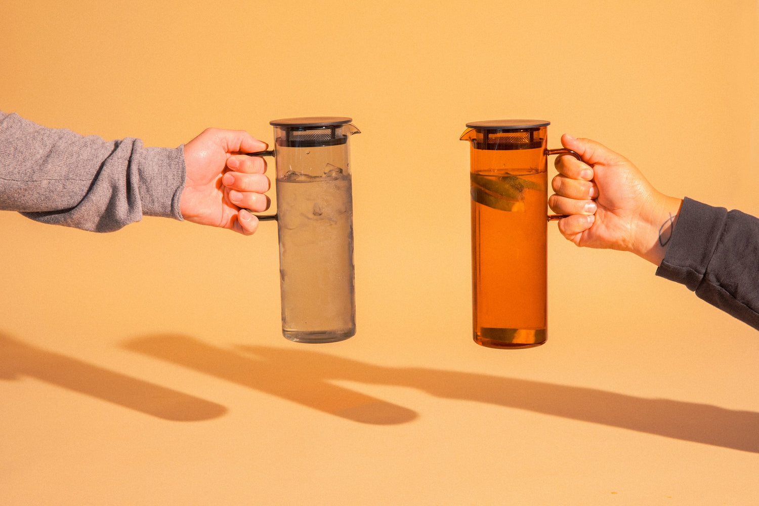 Two cylindrical transparent one gray color glass  and one amber colored pitcher with a fluted spout and rectangular handle with a black colored filtered lid. set on a a light orange background background. Held by 2 different hand