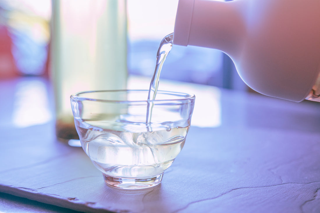 Close up of pouring cold brew tea from a tall glass container with pale grey rubber wine bottle shaped top into a honey-comb shaped short glass tea cup.