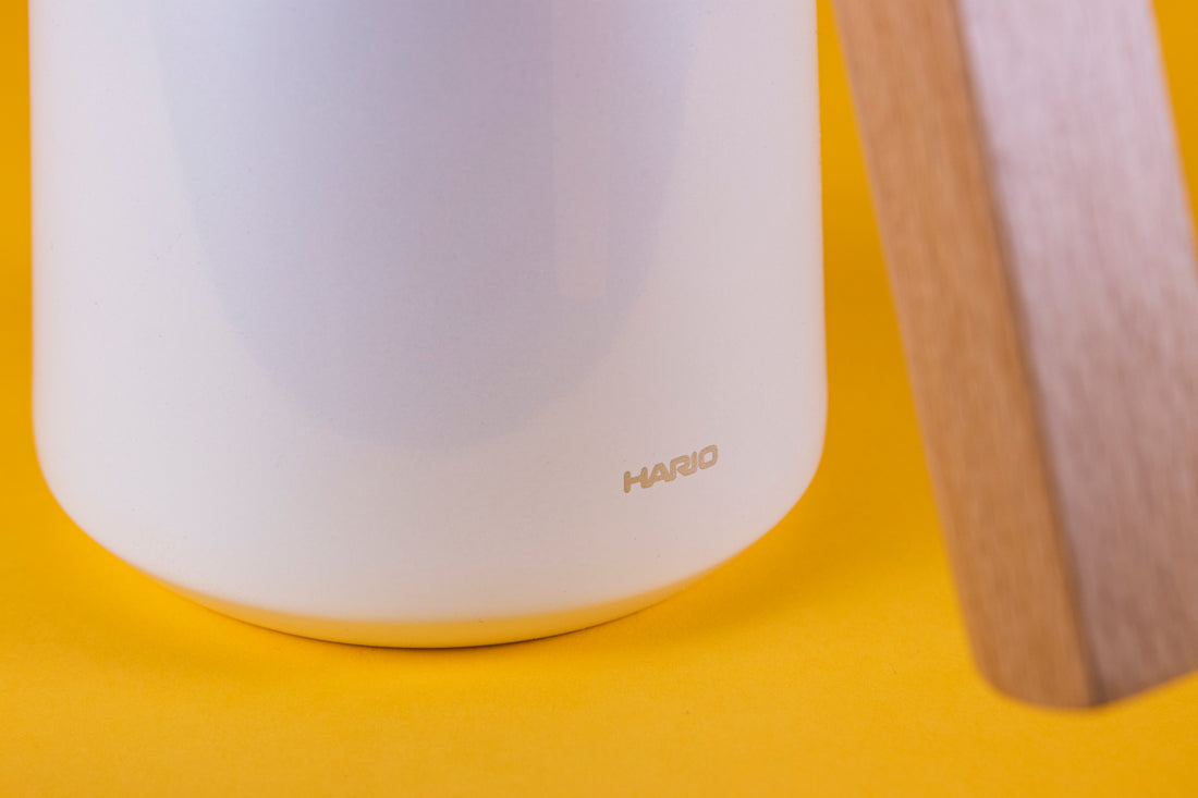 Close up of the Hario logo near the bottom of a white stainless steel thermal serving carafe with wood handle and stainless steel lid set against a yellow background.